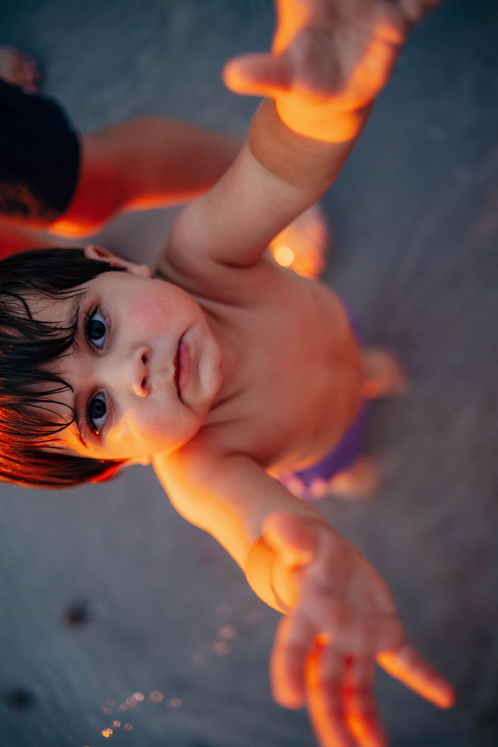 Overhead view of a shirtless toddler with dark hair reaching both arms toward the camera on a sandy beach in warm golden sunset light.