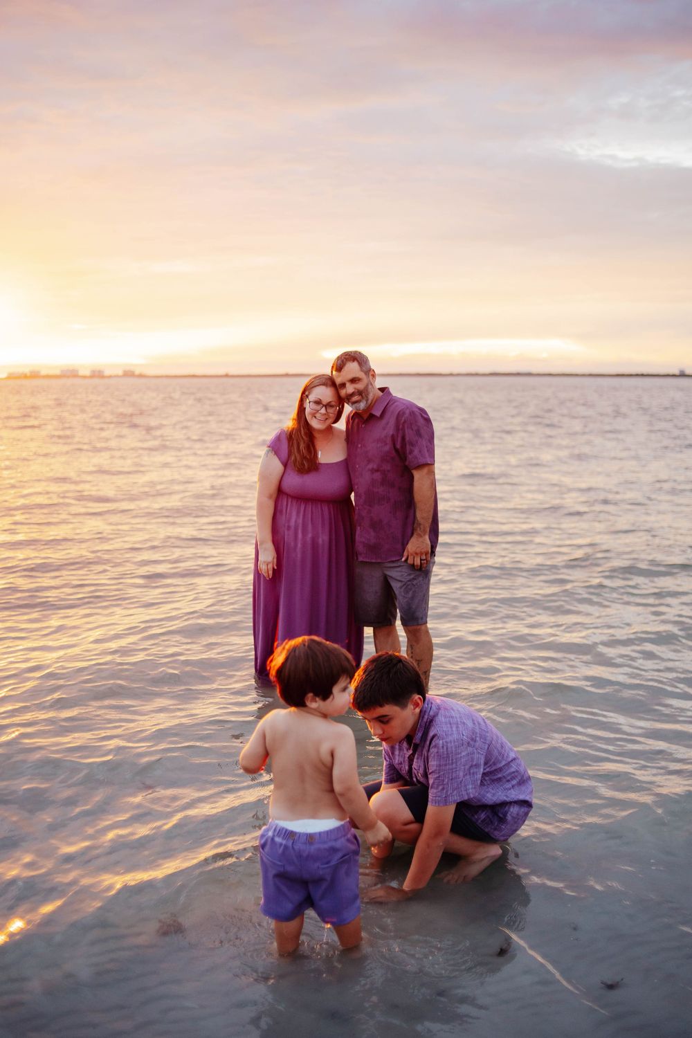 Family in coordinated purple outfits stands in shallow beach water at sunset while two young boys play in the sand.