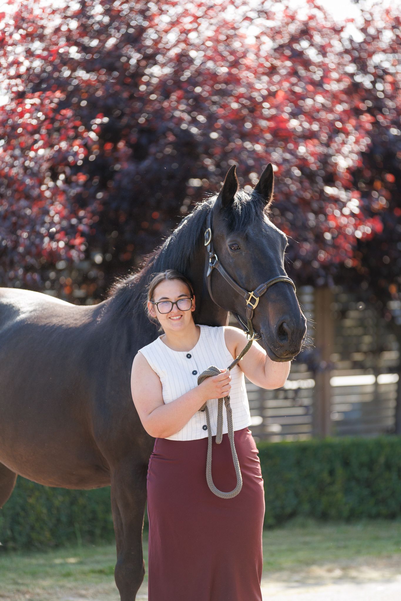 Woman in a white sleeveless top and maroon trousers sits beside a dark brown horse in a bridle, gently touching its muzzle outdoors.