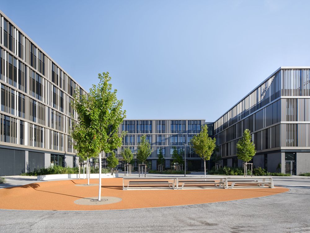 Modern glass and metal office buildings surround a courtyard with young trees, wooden benches and an orange paved plaza under a clear sky.