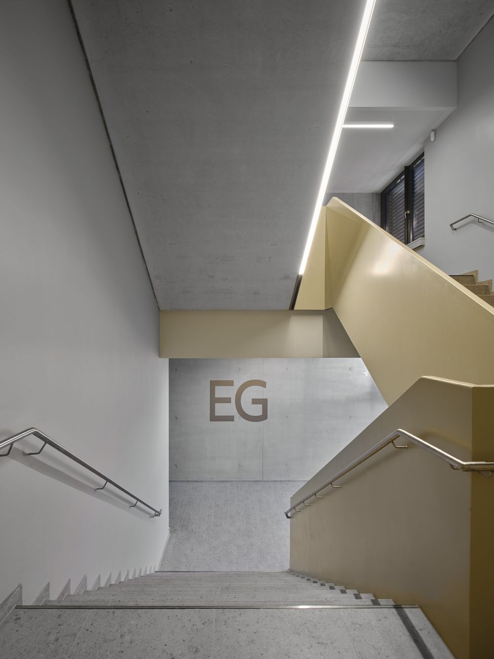 Modern concrete stairwell with terrazzo steps, golden railings, linear ceiling lighting and an EG ground floor sign on the wall.