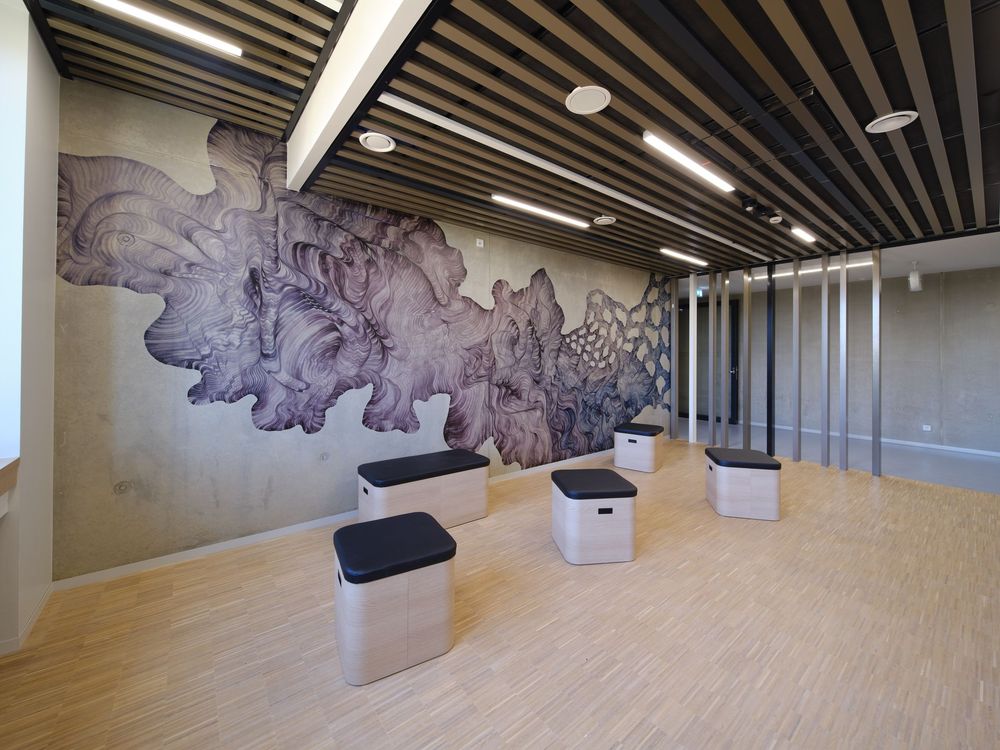 Modern lounge with a large intricate black-and-white mural, wood slat ceiling, light wood floors, and square gray and black stool seats.