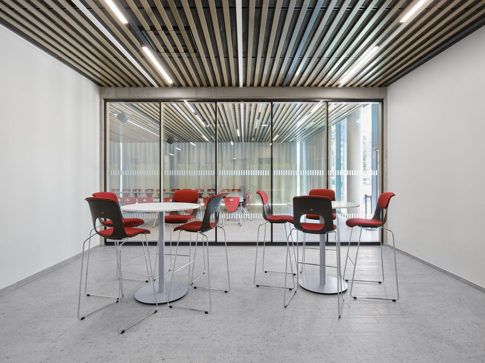 Modern breakout area with round high tables, red and black barstools, a slatted wood ceiling and floor-to-ceiling glass wall.