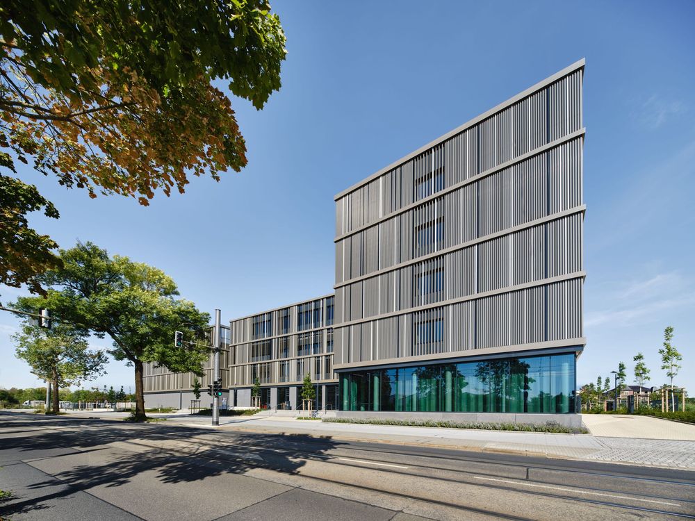 Modern multi-story office building with a slanted glass and metal facade beside a tree-lined street under a clear blue sky.