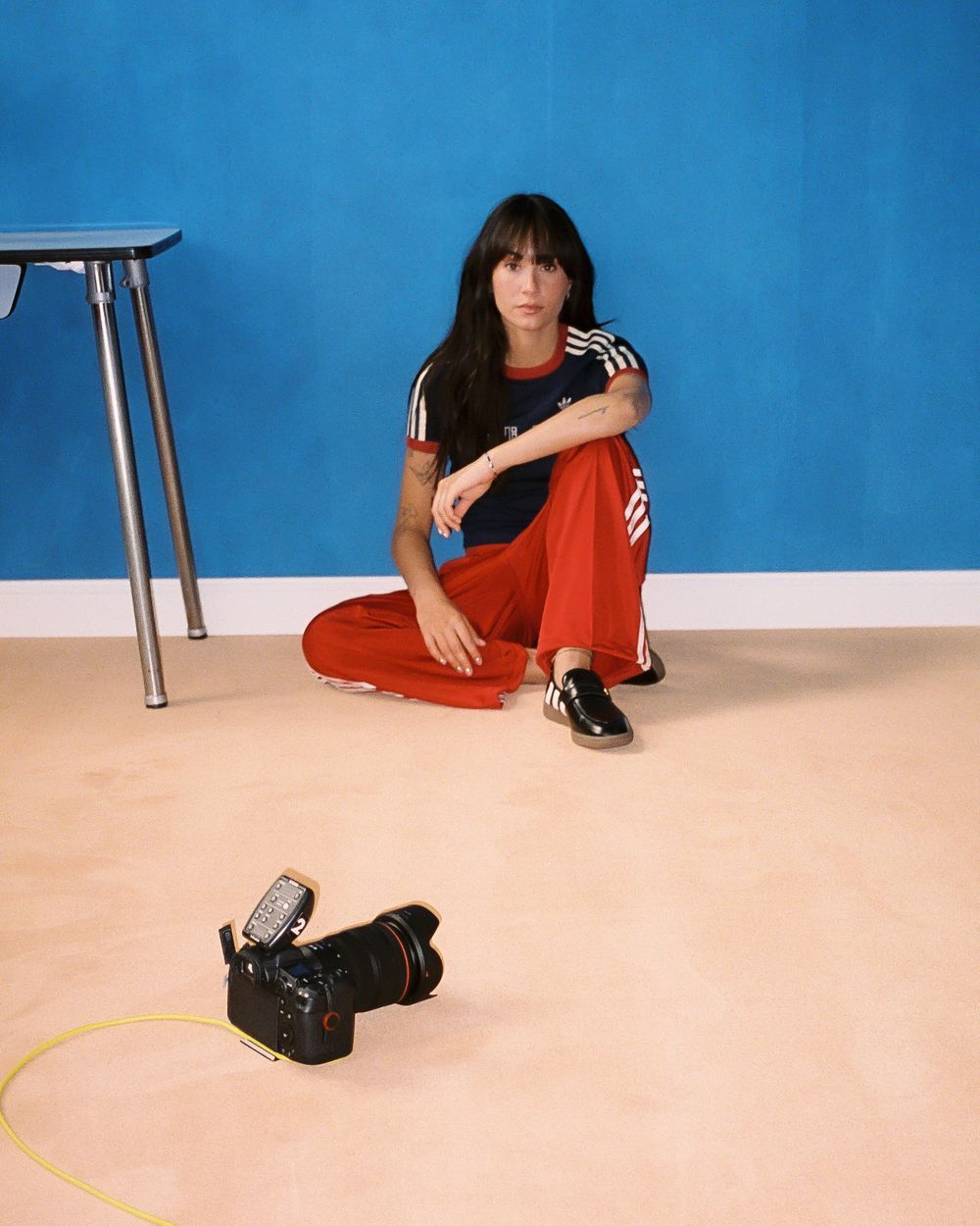 Woman in red Adidas tracksuit sits on floor against blue wall with DSLR camera nearby.