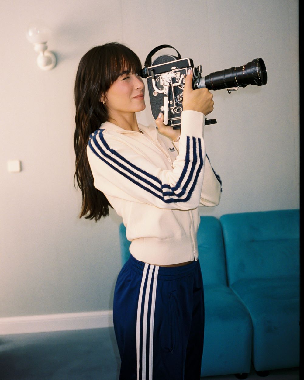 Woman in white Adidas tracksuit holds vintage film camera to her eye, standing beside a teal sofa.
