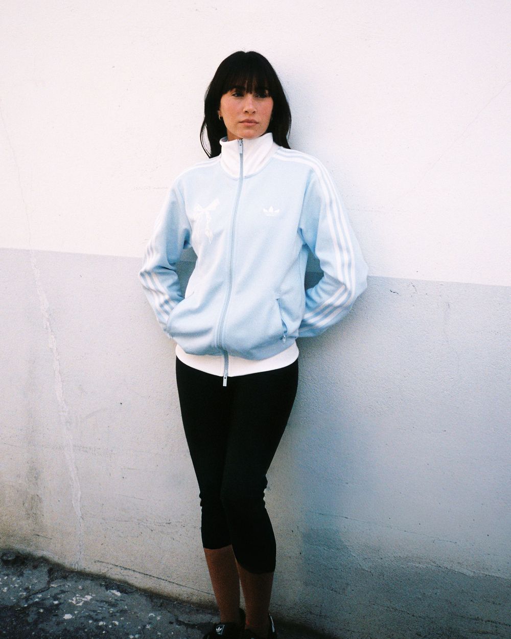 Woman in light blue Adidas track jacket and black leggings leaning against a white wall outdoors.