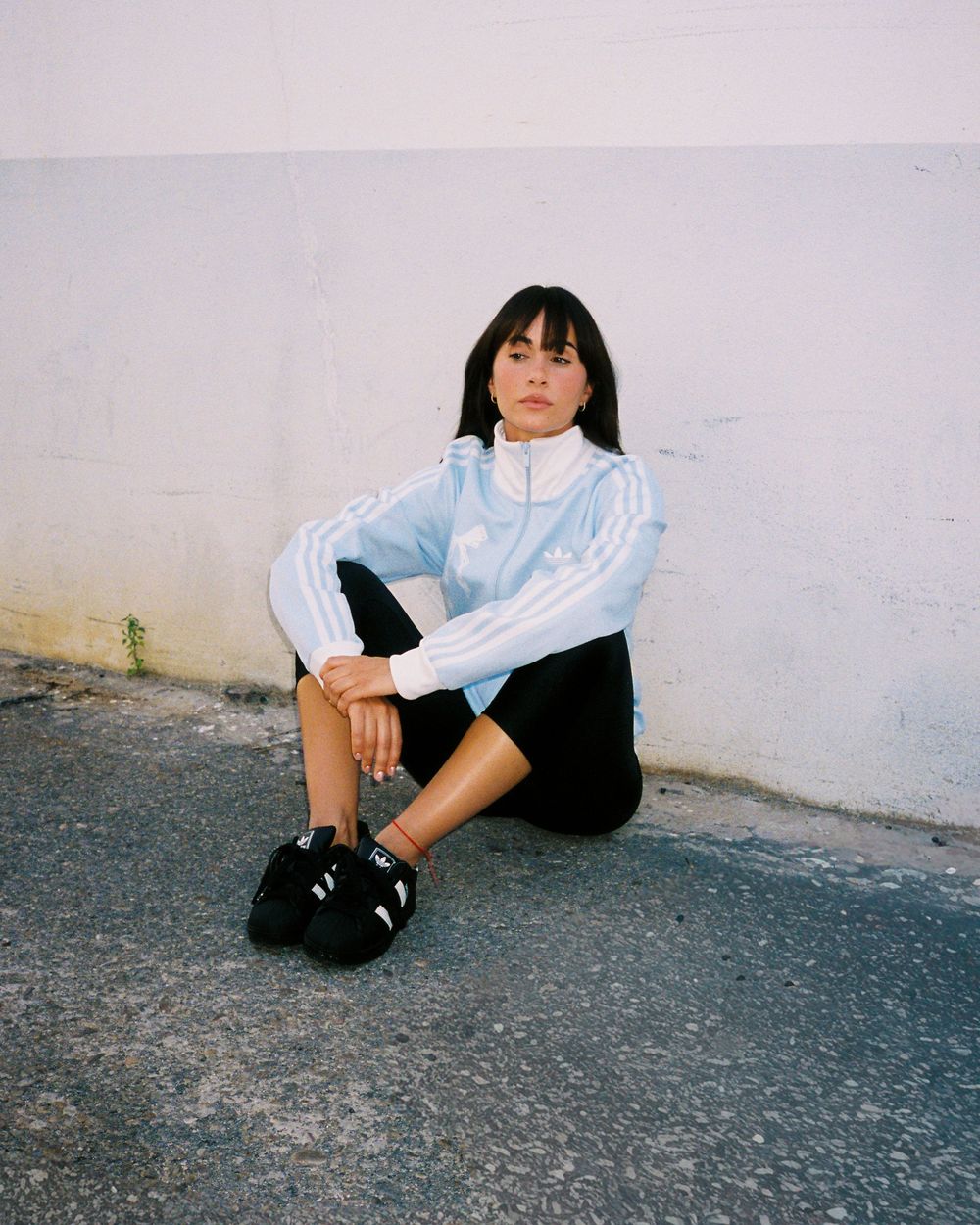 Young woman in Adidas light blue track jacket and black shorts sits against a white wall on pavement.