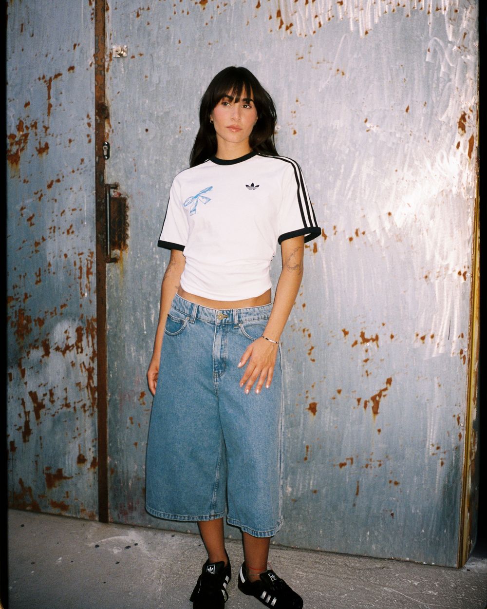 Woman wearing white Adidas crop tee with black stripes, wide-leg denim shorts, and black sneakers, posing against rusty metal wall.