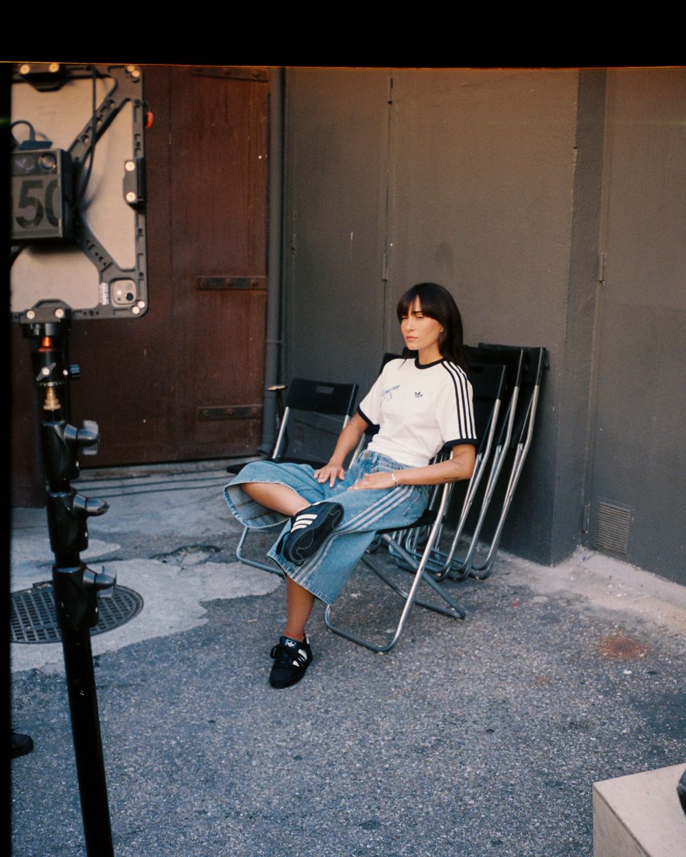Person sitting on a folding chair outdoors wearing an Adidas white tee, denim skirt, and black sneakers near a metal door.