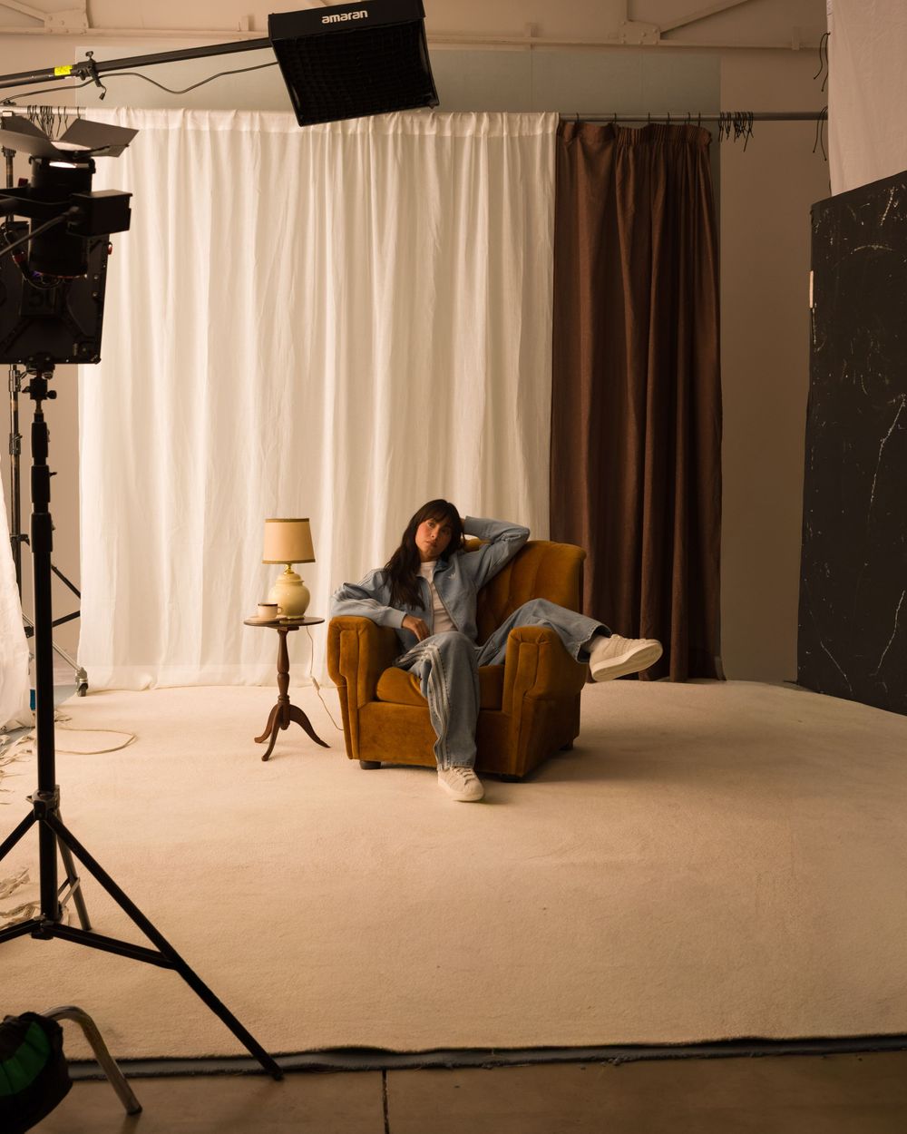 Woman in blue denim outfit sits in gold armchair on white carpet in a professional photography studio setup.