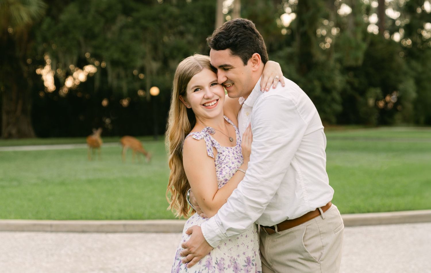 Smiling couple embracing on a garden path with deer grazing behind them during a Jekyll Island engagement session.