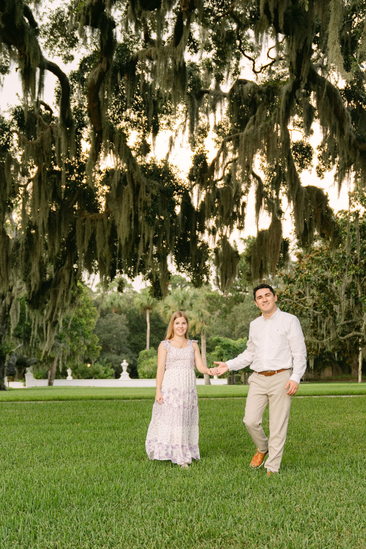 Couple walking hand in hand across a green lawn beneath oak trees during their Jekyll Island engagement session.