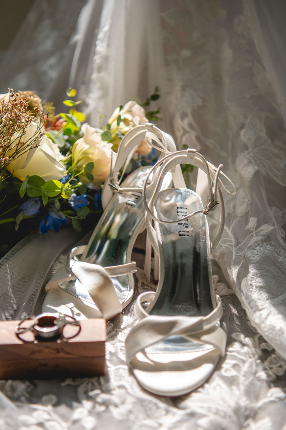 Elegant white bridal heels displayed on lace fabric beside a floral bouquet and wooden ring box.