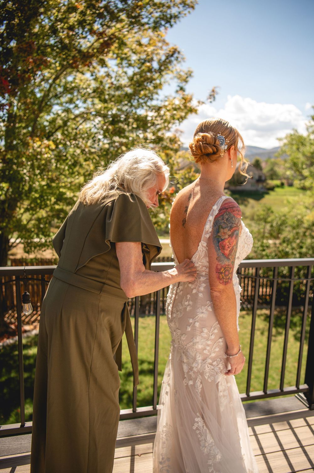 Older woman helping bride button her lace wedding dress on a sunny outdoor balcony, bride has floral arm tattoo.