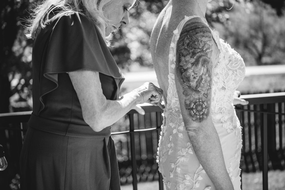 Black and white photo of bride in lace gown with tattoo sleeve, assisted by woman in dark dress at outdoor wedding.