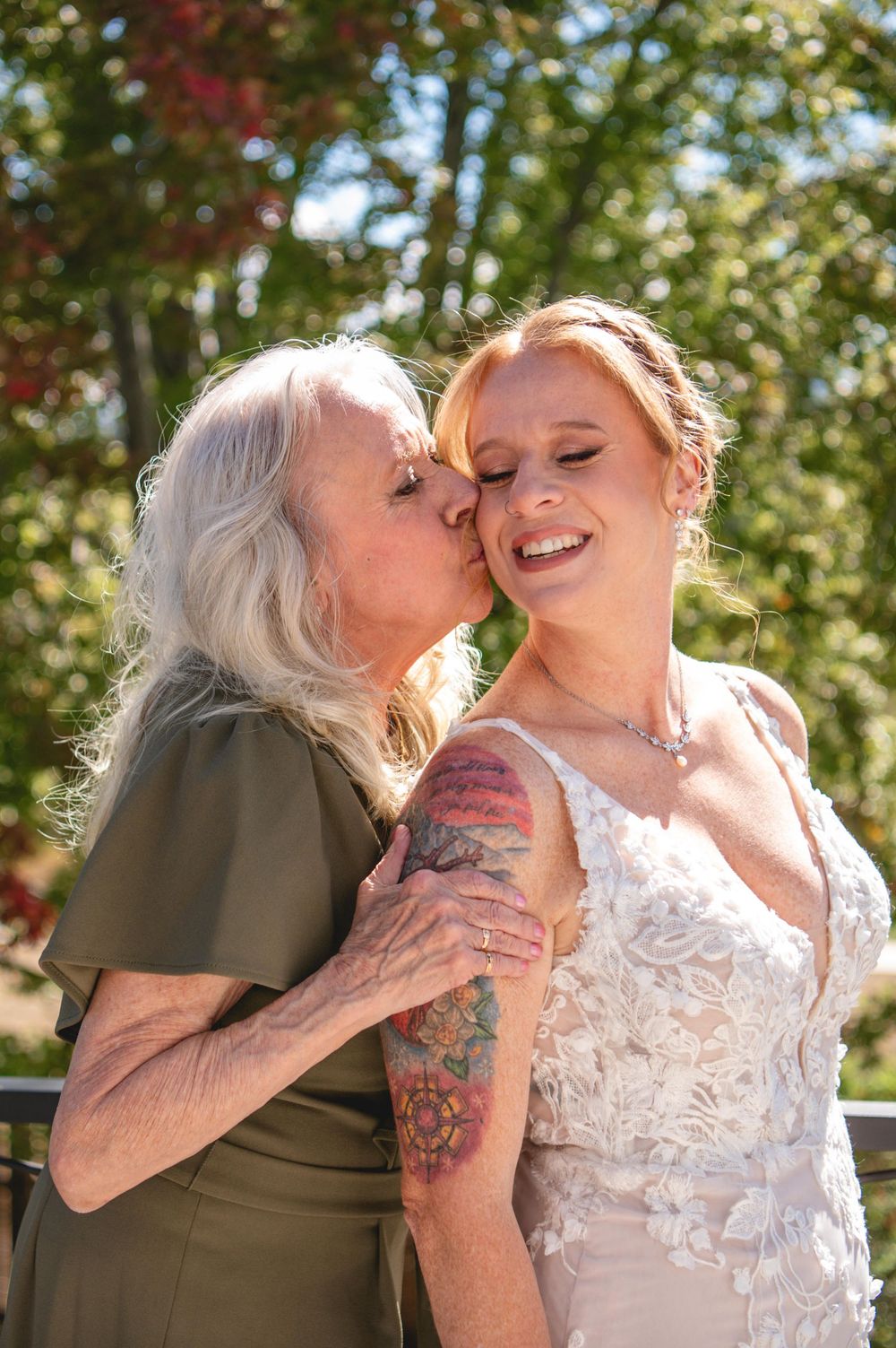 Elderly woman kissing the cheek of a smiling bride in a lace wedding dress with a floral arm tattoo outdoors.