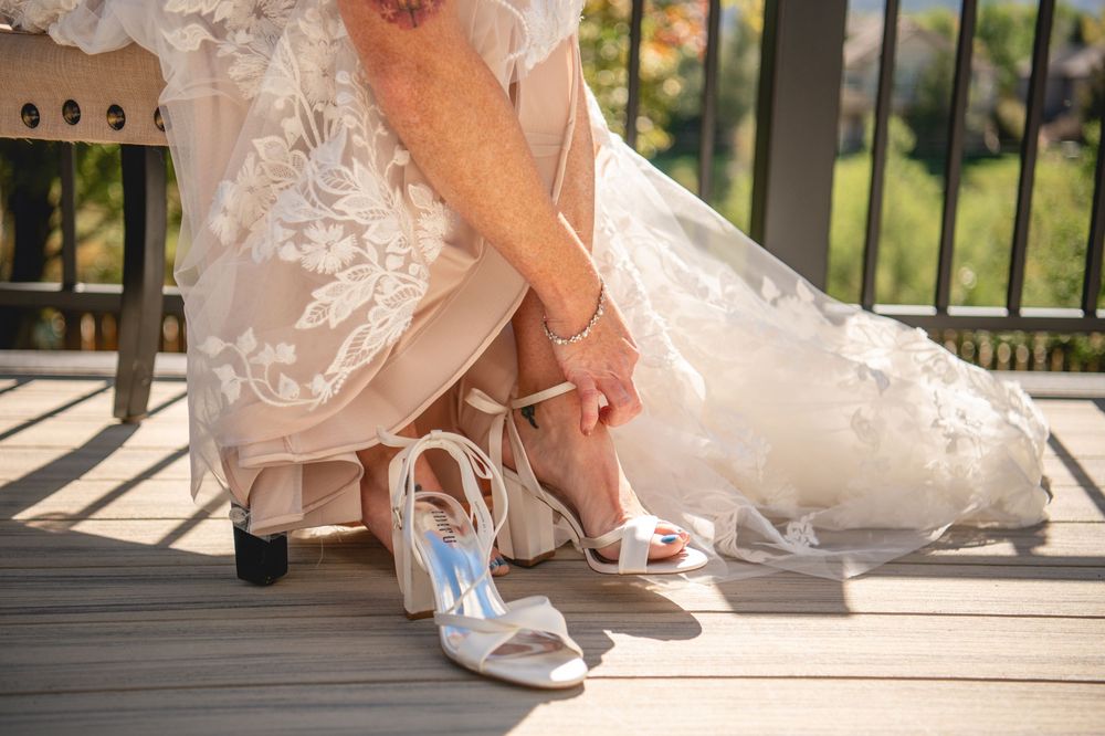 Bride putting on white strappy wedding heels on a wooden deck, with lace bridal gown draped around her feet.