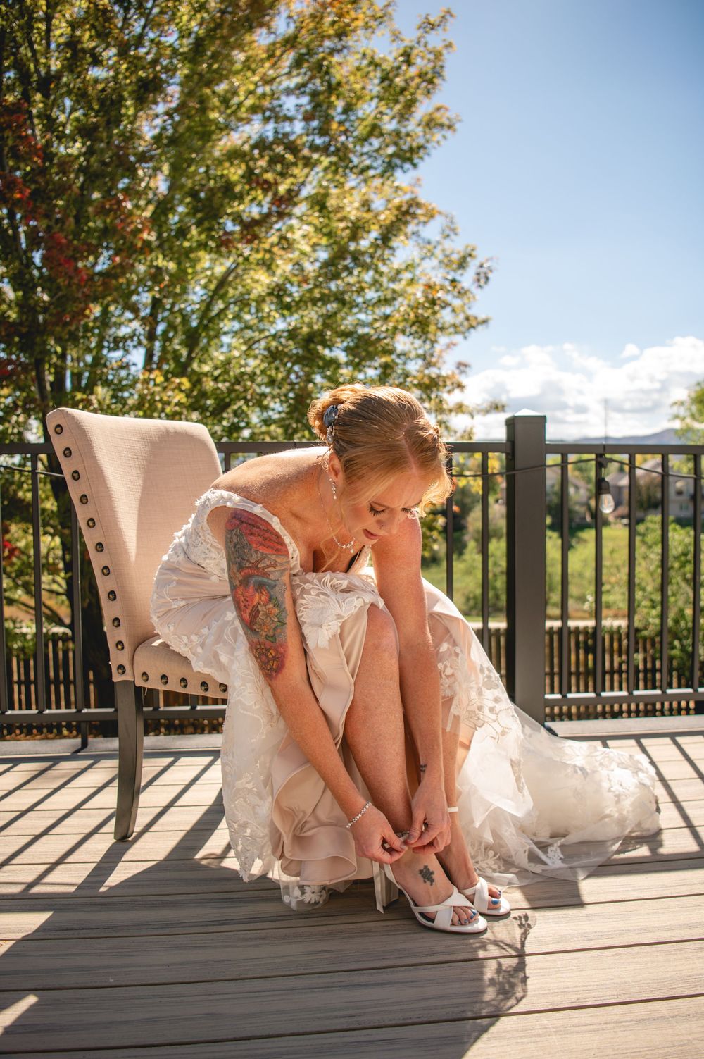Bride in white lace wedding dress adjusting her heels on outdoor deck chair, tattoos visible, surrounded by green trees.