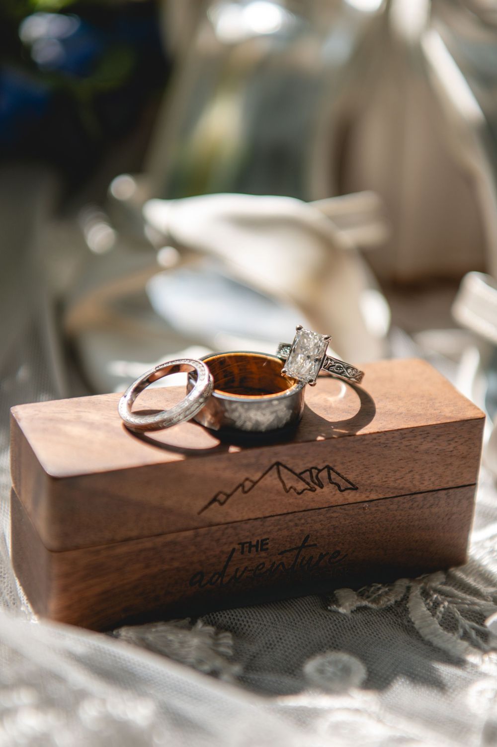 Three wedding rings displayed on a rustic wooden engraved ring box with mountain design and cursive text reading The Adventure.
