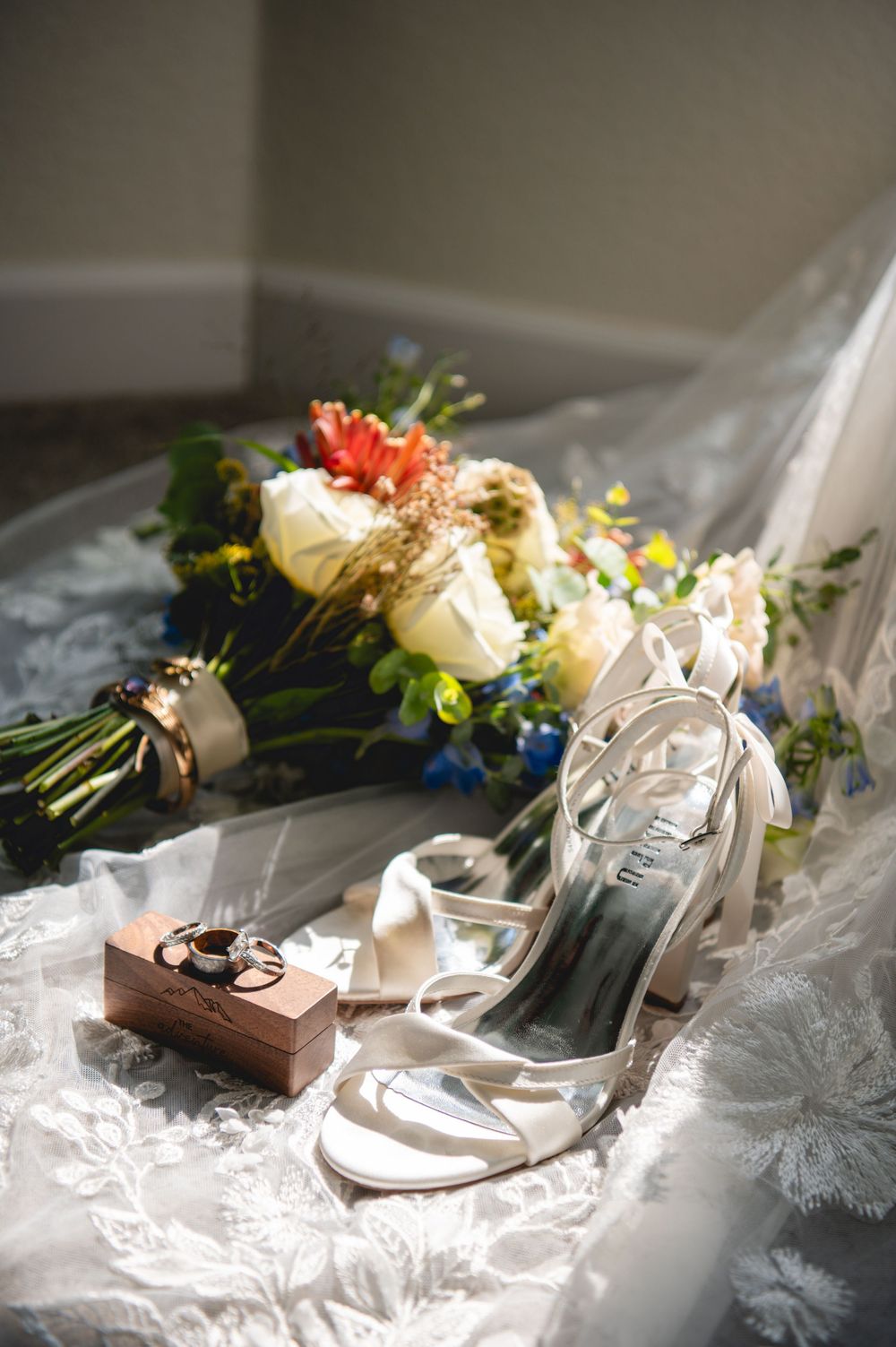 White bridal heels, wedding rings in wooden box, and colorful floral bouquet arranged on lace veil in soft natural light.