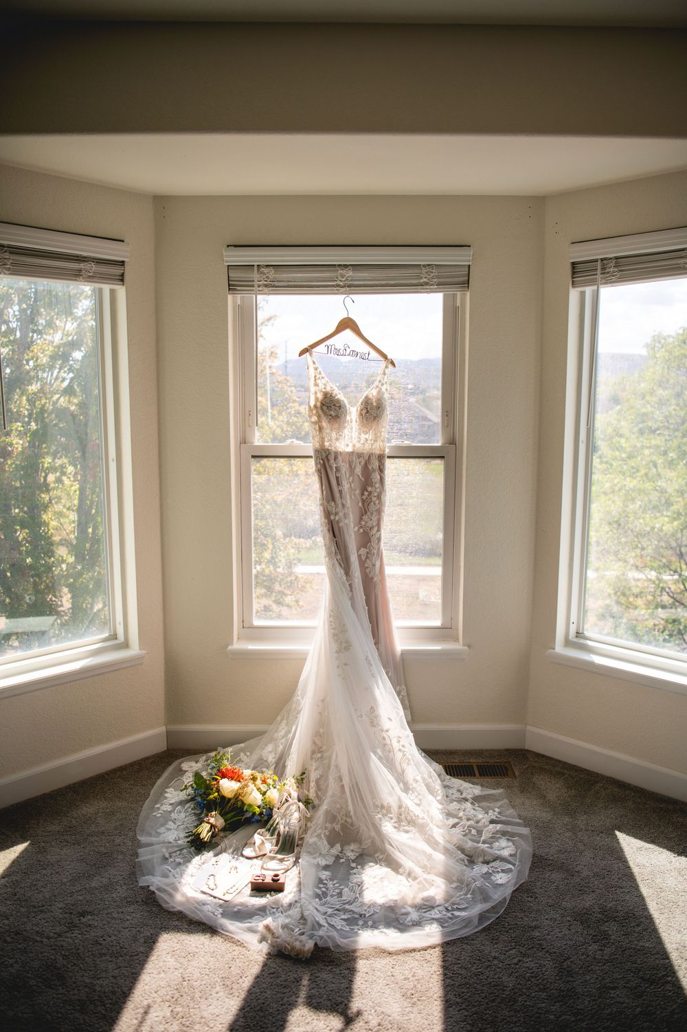 Elegant lace wedding dress hanging in a sunlit bay window with a floral bouquet and accessories on the floor.