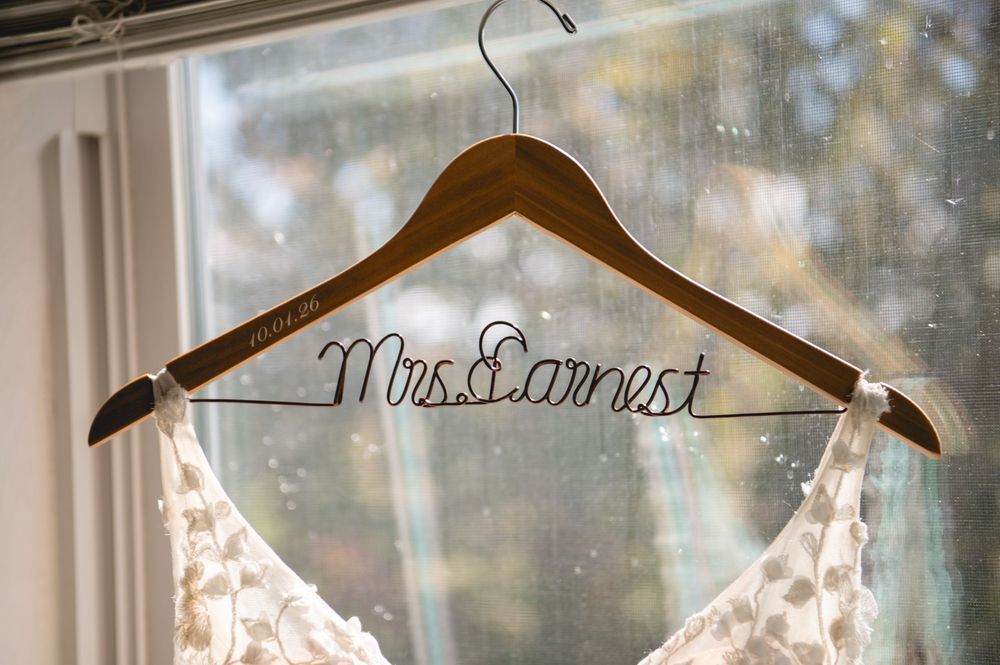 Custom wooden wedding hanger with wire Mrs. Earnest name detail holding a white lace bridal gown by a window.