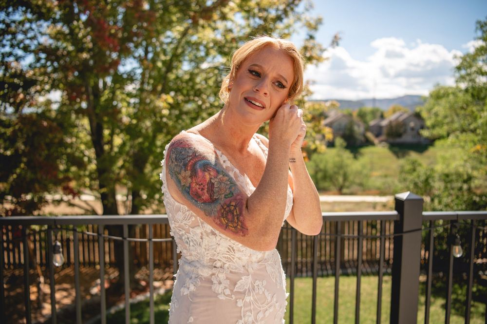 Tattooed bride in lace wedding dress adjusting earring on outdoor balcony surrounded by lush green trees.