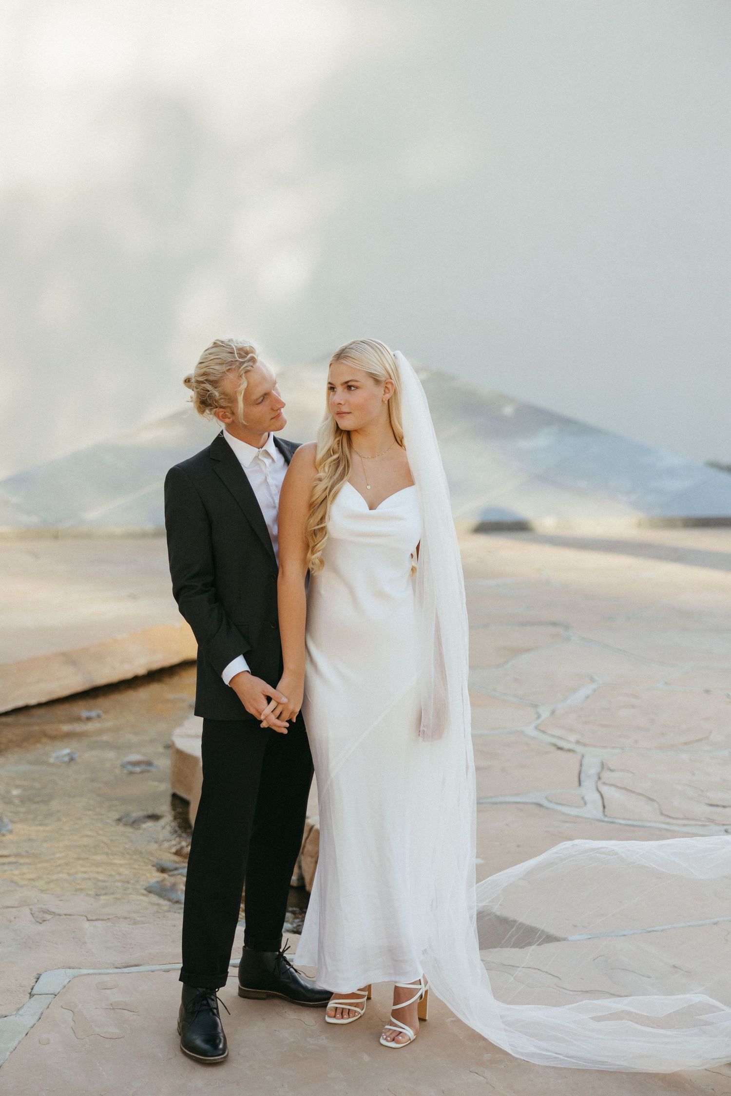 Bride and groom share a romantic moment outdoors, she holds a red and white cascading bouquet, both in elegant wedding attire.
