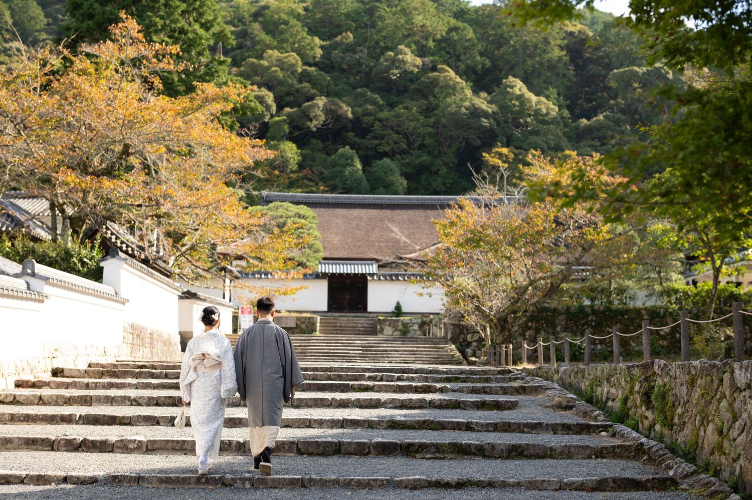 Couple in traditional Japanese kimono walking up stone steps toward a temple surrounded by autumn foliage.