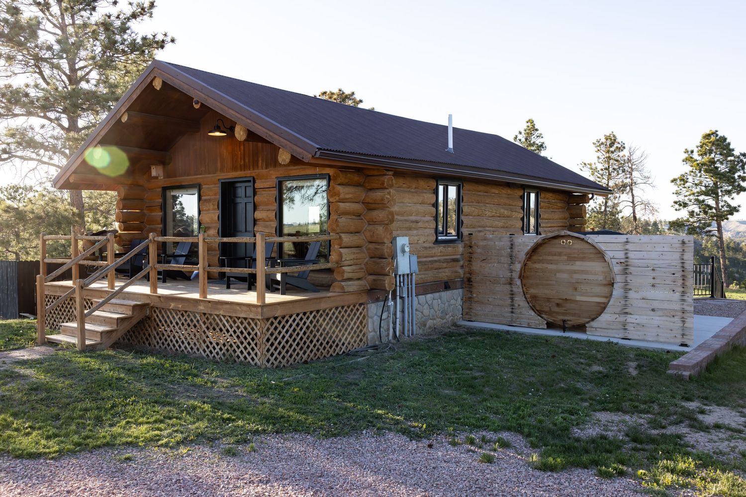 Rustic log cabin with a covered wooden porch, stairs, and a round barrel-style sauna surrounded by pine trees and a grassy yard.
