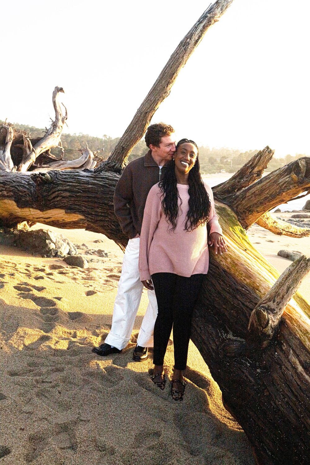 Couple smiling and embracing by driftwood logs on a sandy beach at sunset.