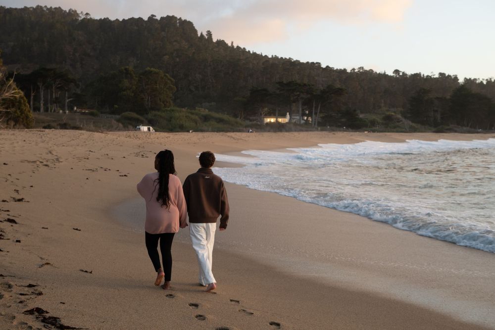 Couple walking hand in hand along a sandy beach at sunset with ocean waves and tree-lined hills in background.