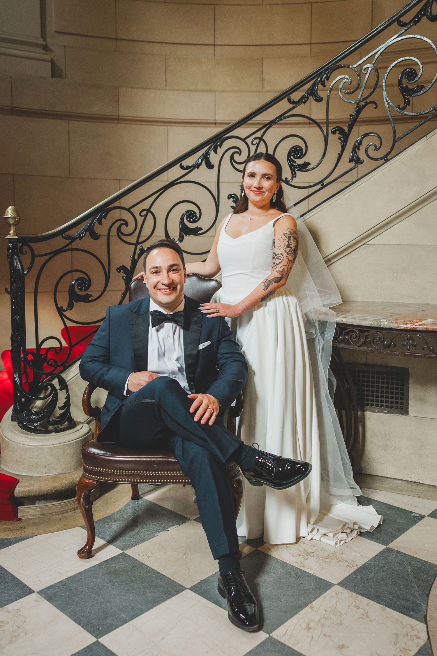 Bride and groom posing on ornate staircase, groom seated in vintage chair wearing tuxedo, bride in white gown with veil and tattoo.