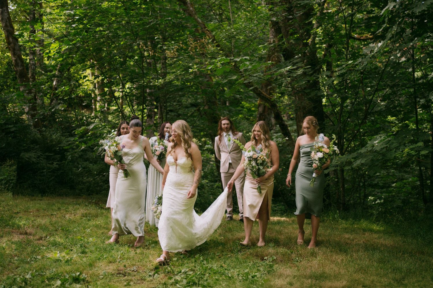 Bridesmaids in colorful dresses help a bride in a white gown and veil near a floral arch at an outdoor wedding.