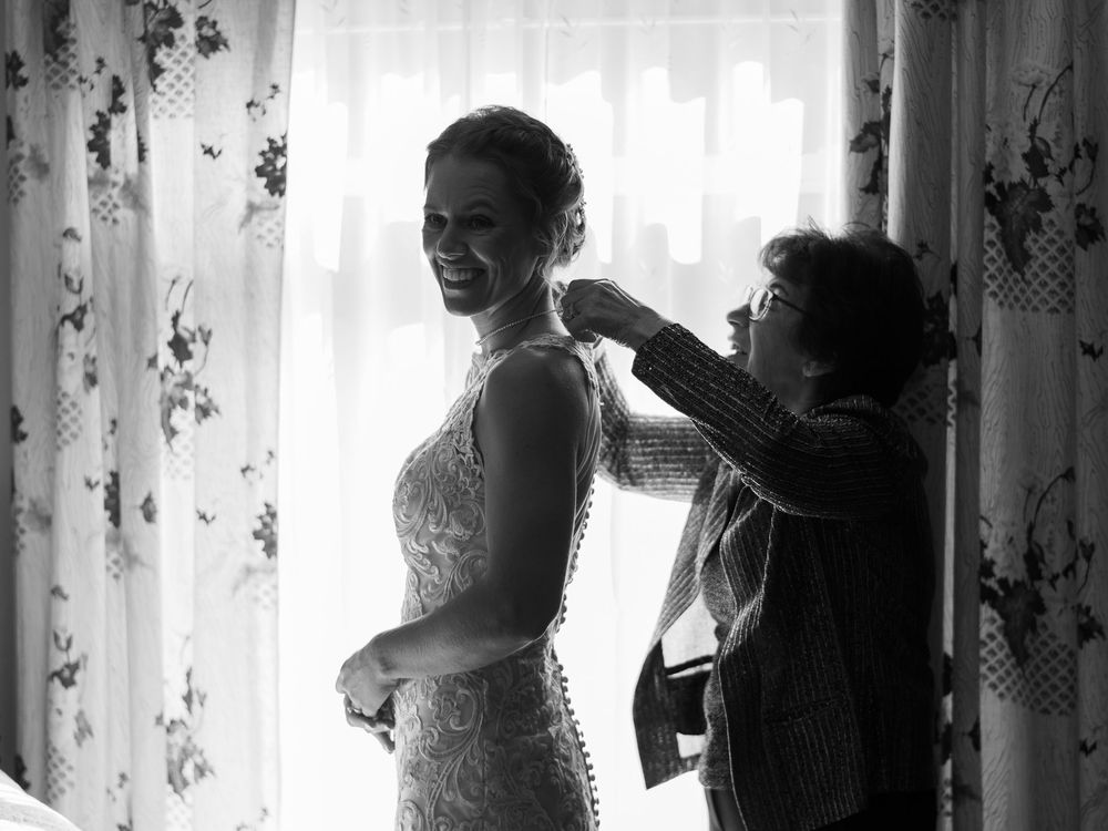 Black and white photo of a bride smiling as a woman helps button her lace wedding dress near a curtained window.