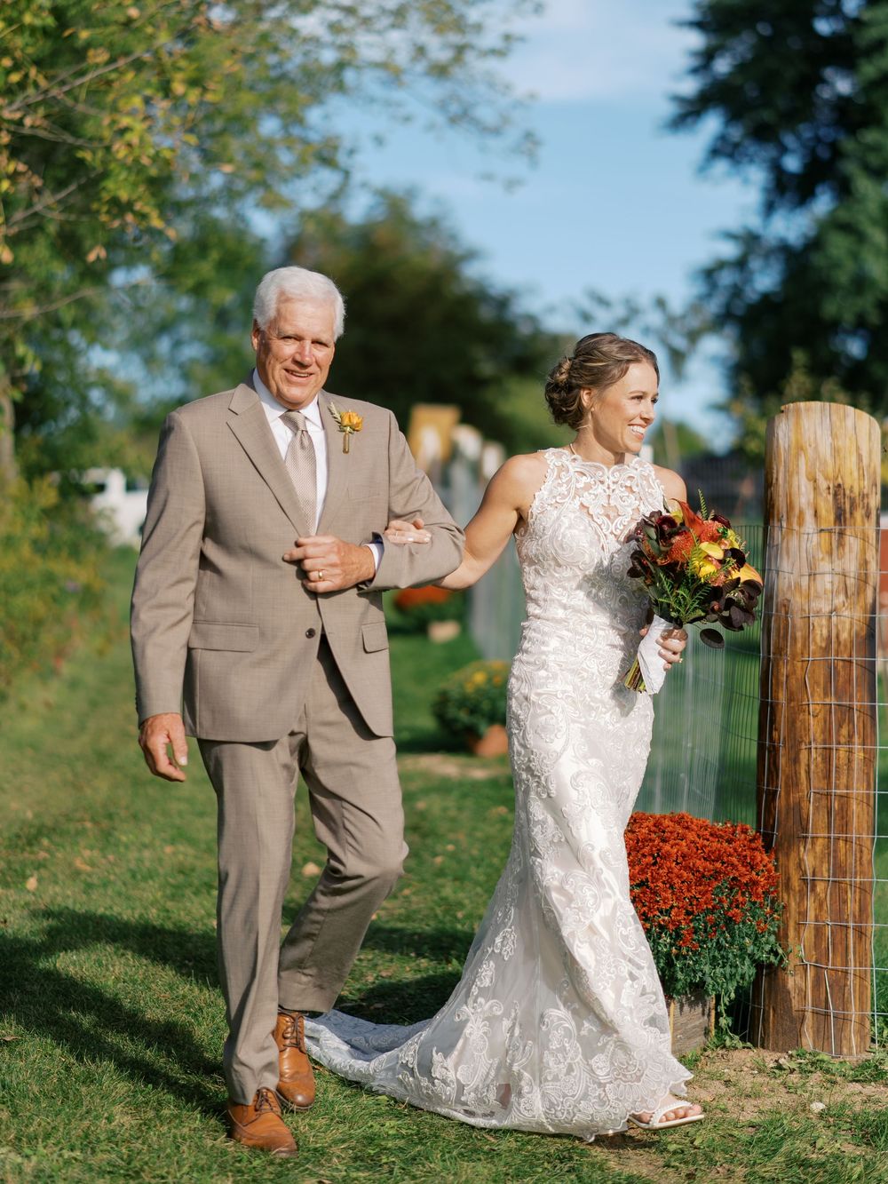 Bride in lace gown holding fall bouquet walks down aisle with father in tan suit at outdoor autumn wedding.
