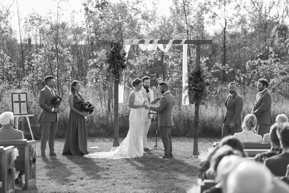 Black and white outdoor wedding ceremony with couple at floral arch, wedding party, and seated guests in a natural wooded setting.
