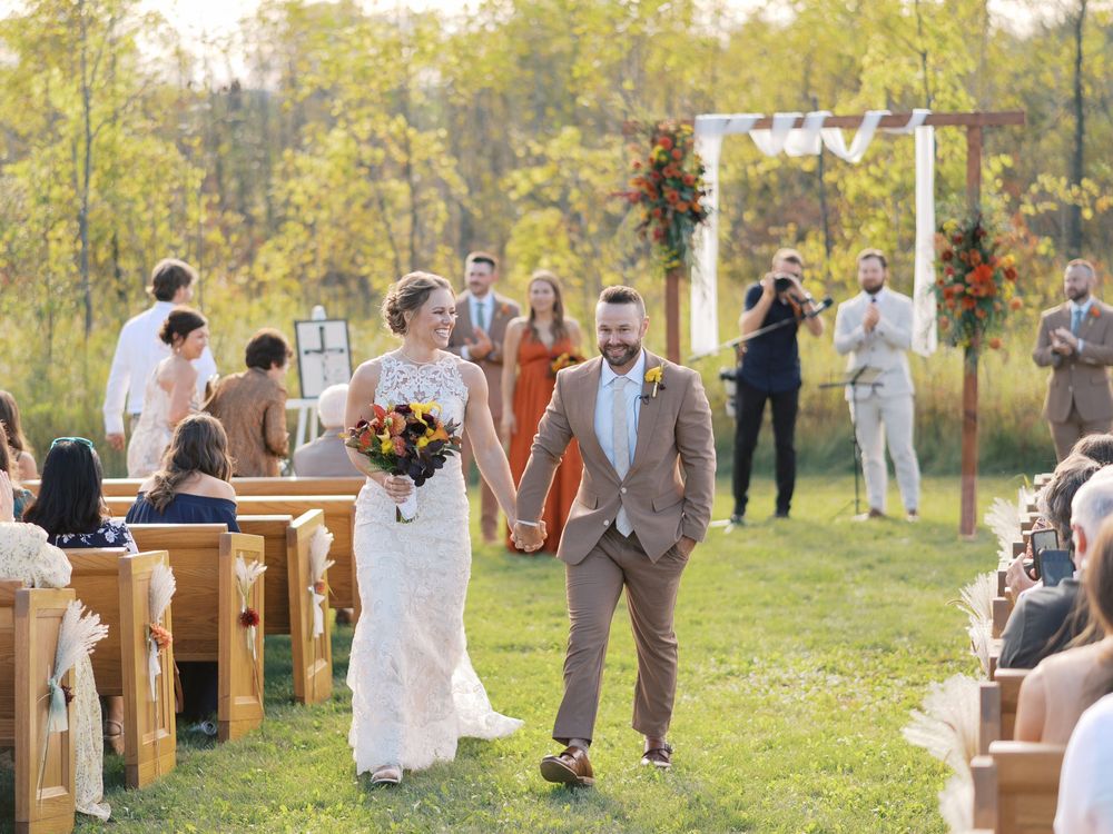Bride and groom walk down outdoor wedding aisle holding hands, surrounded by guests on wooden pews in a lush green field.