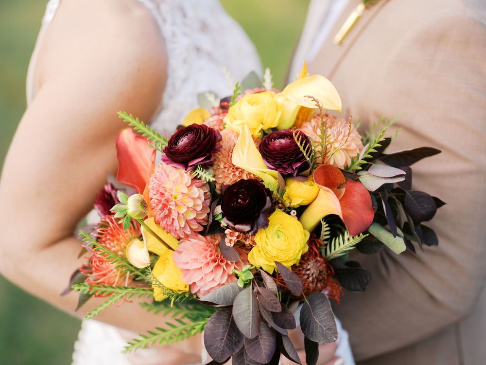 Bride holding a vibrant wedding bouquet with yellow, coral, and burgundy flowers including dahlias and calla lilies.