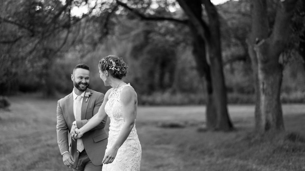 Black and white photo of a smiling groom holding hands with his bride in a lace dress outdoors.