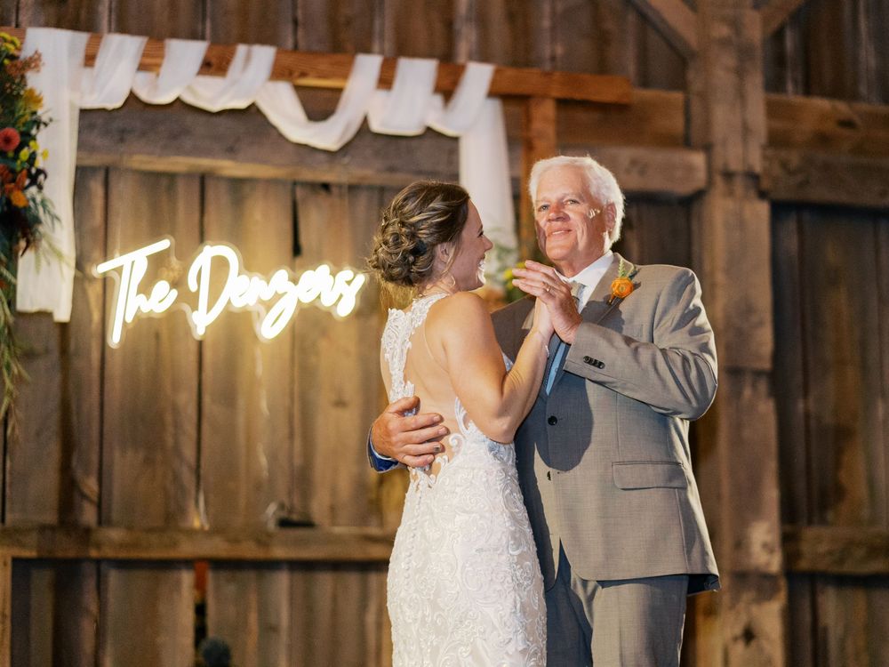 Bride and father share first dance in rustic barn venue with glowing neon sign reading The Danzers.