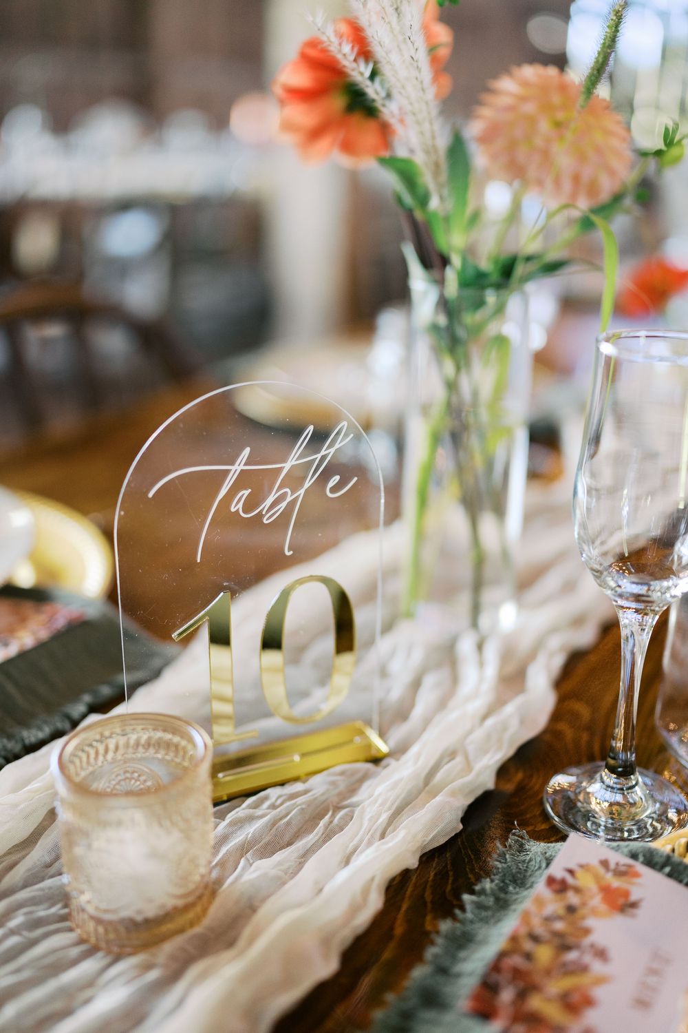 Elegant wedding table number 10 sign on acrylic with gold stand, floral centerpiece, and glassware on rustic table.