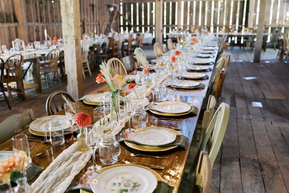 Rustic barn wedding reception with long wooden tables set with elegant plates, glassware, and floral centerpieces.
