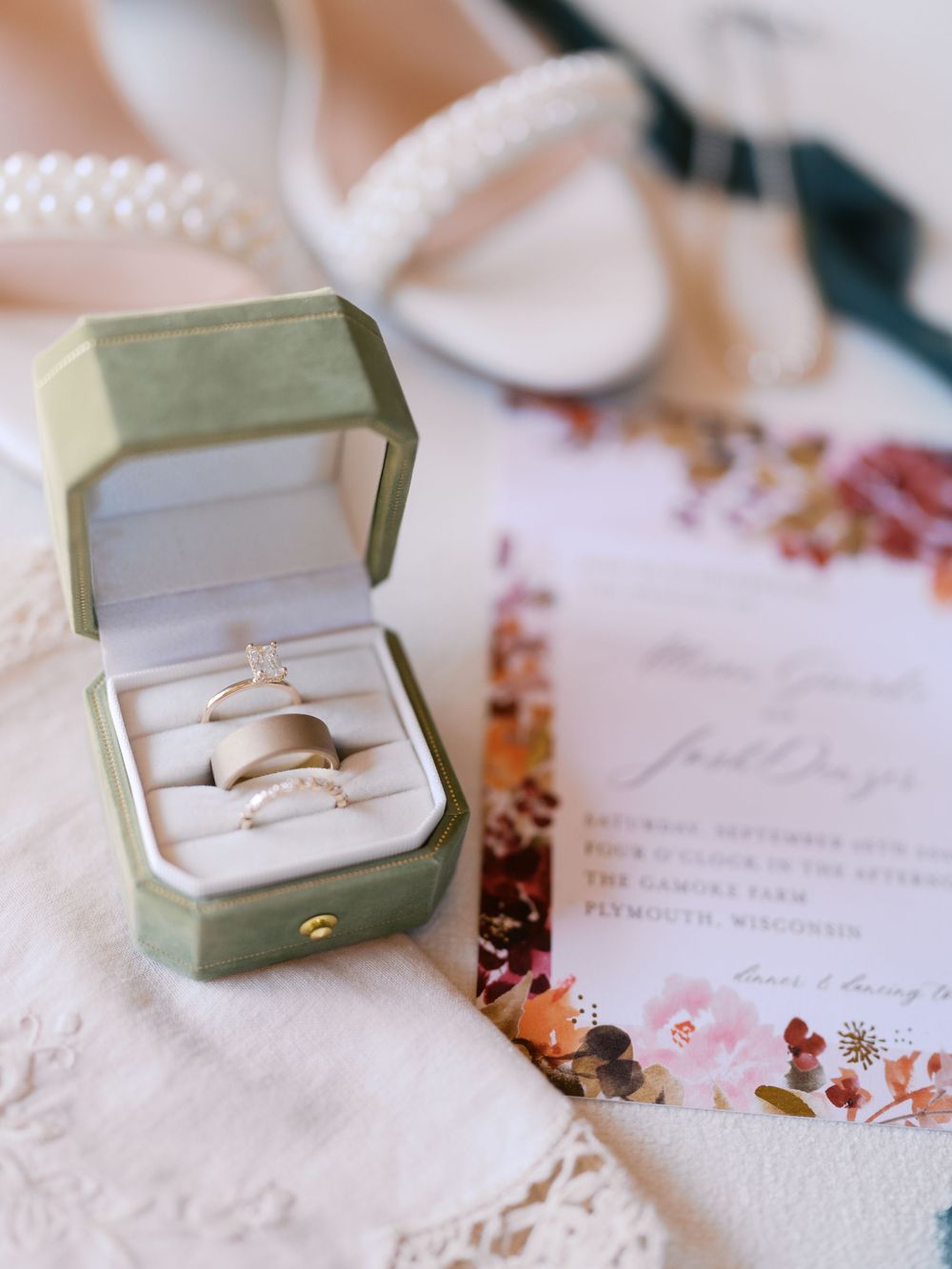 Green velvet ring box displaying wedding bands and engagement ring beside floral invitation and pearl necklace.