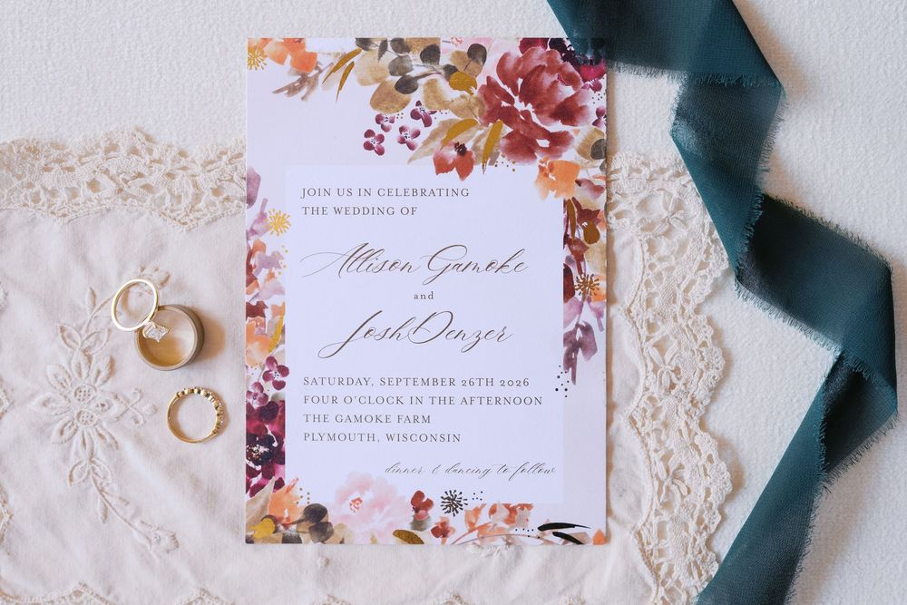 Floral wedding invitation with autumn blooms, gold rings, and teal ribbon on lace fabric for a fall wedding.