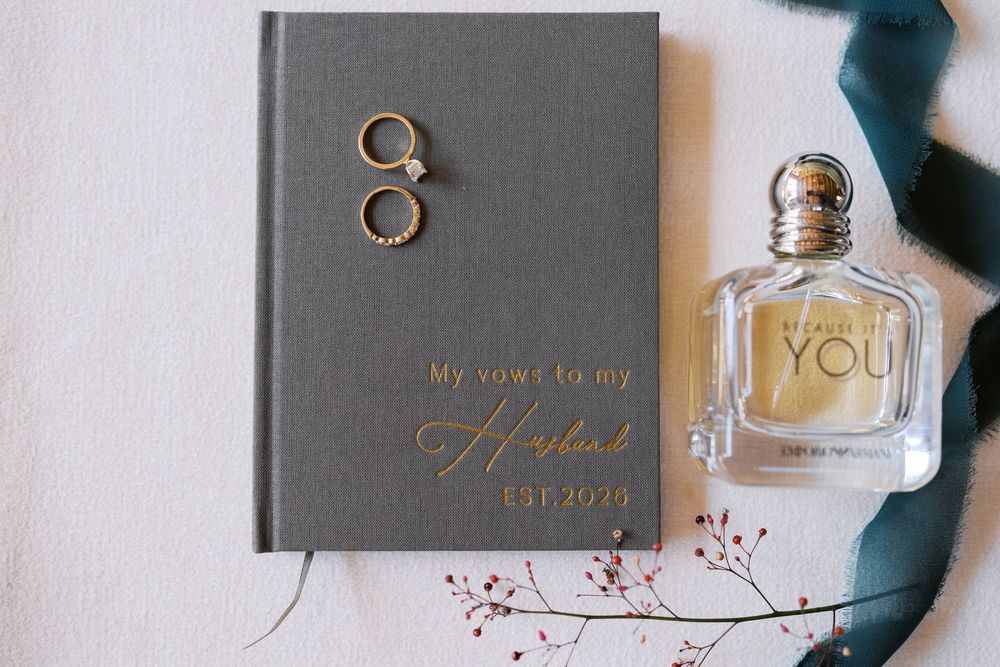 Gray vow book with gold script, wedding rings, Emporio Armani perfume, and teal ribbon on white background.