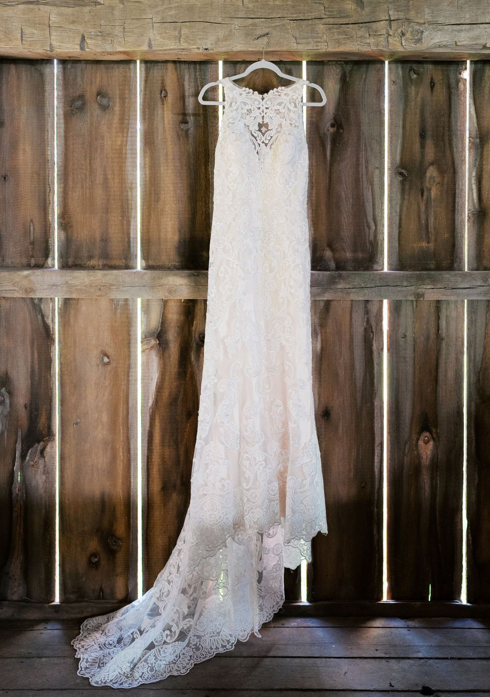 Elegant lace wedding dress with a long train hanging on a rustic wooden barn wall.