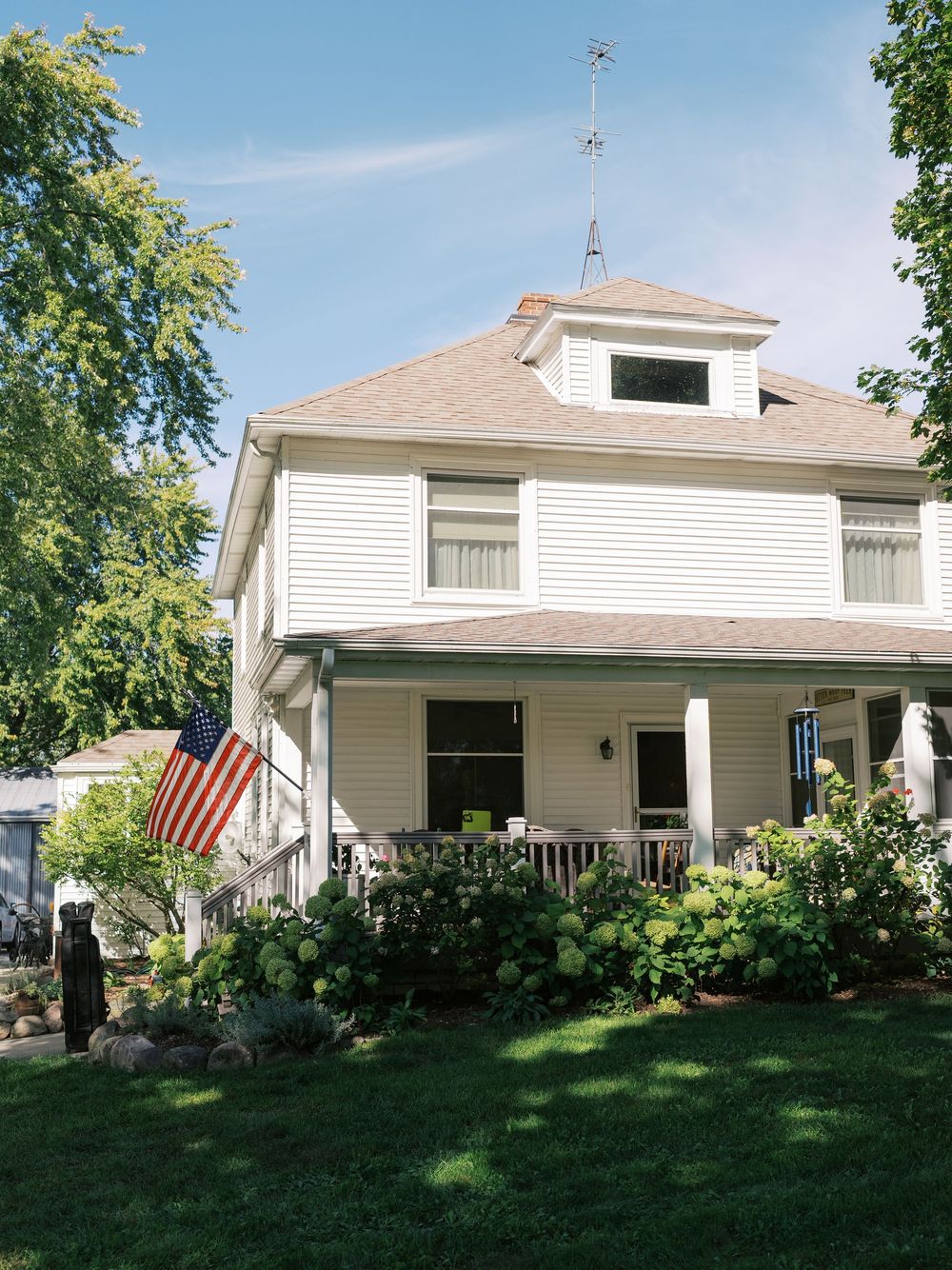 White two-story American home with wrap-around porch, US flag, lush garden, and blue sky on a sunny day.