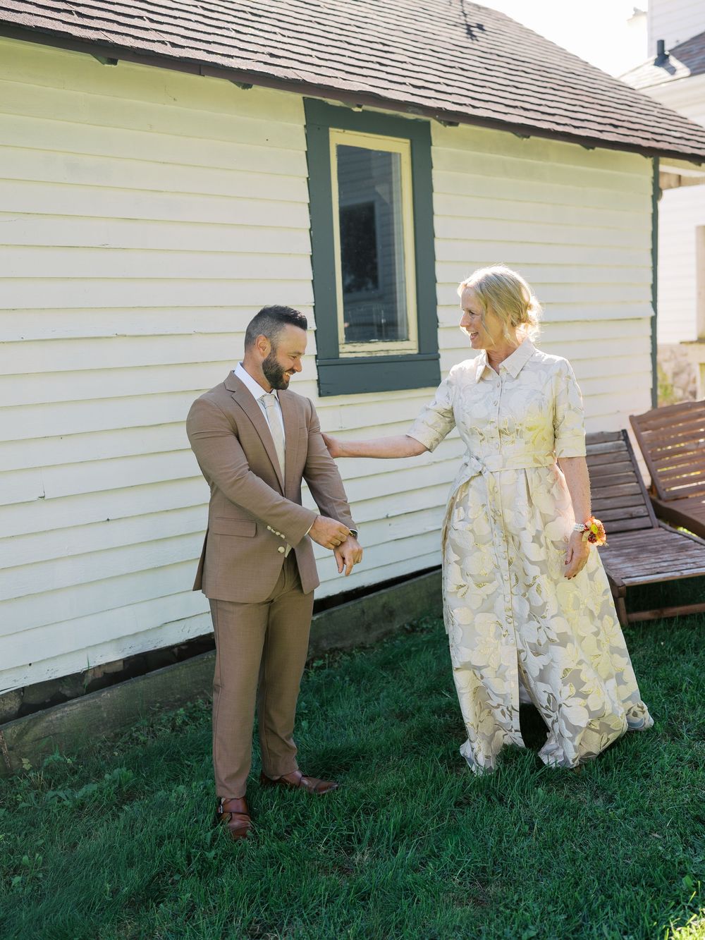 A couple in formal attire dancing on green grass beside a white farmhouse on a sunny day.